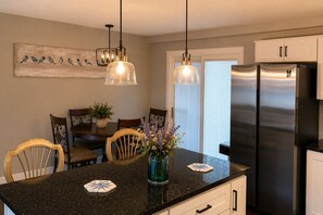 A kitchen with a black granite countertop, a stainless steel refrigerator, two pendant lights, and a dining area with wooden chairs and a table. - Hidden Sapphire Shores - Near Beach + Fire Pit + Family Retreat (Michigan City)