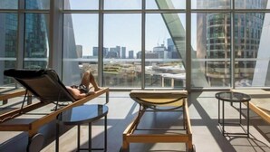 En moderne lounge med store vinduer, en hvilestol, et lille bord og udsigt til bybillede.