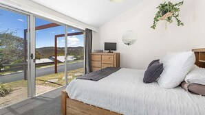 Une chambre avec une grande porte-fenêtre coulissante, un lit avec une literie blanche, une table de chevet en bois, une télévision et une plante suspendue.