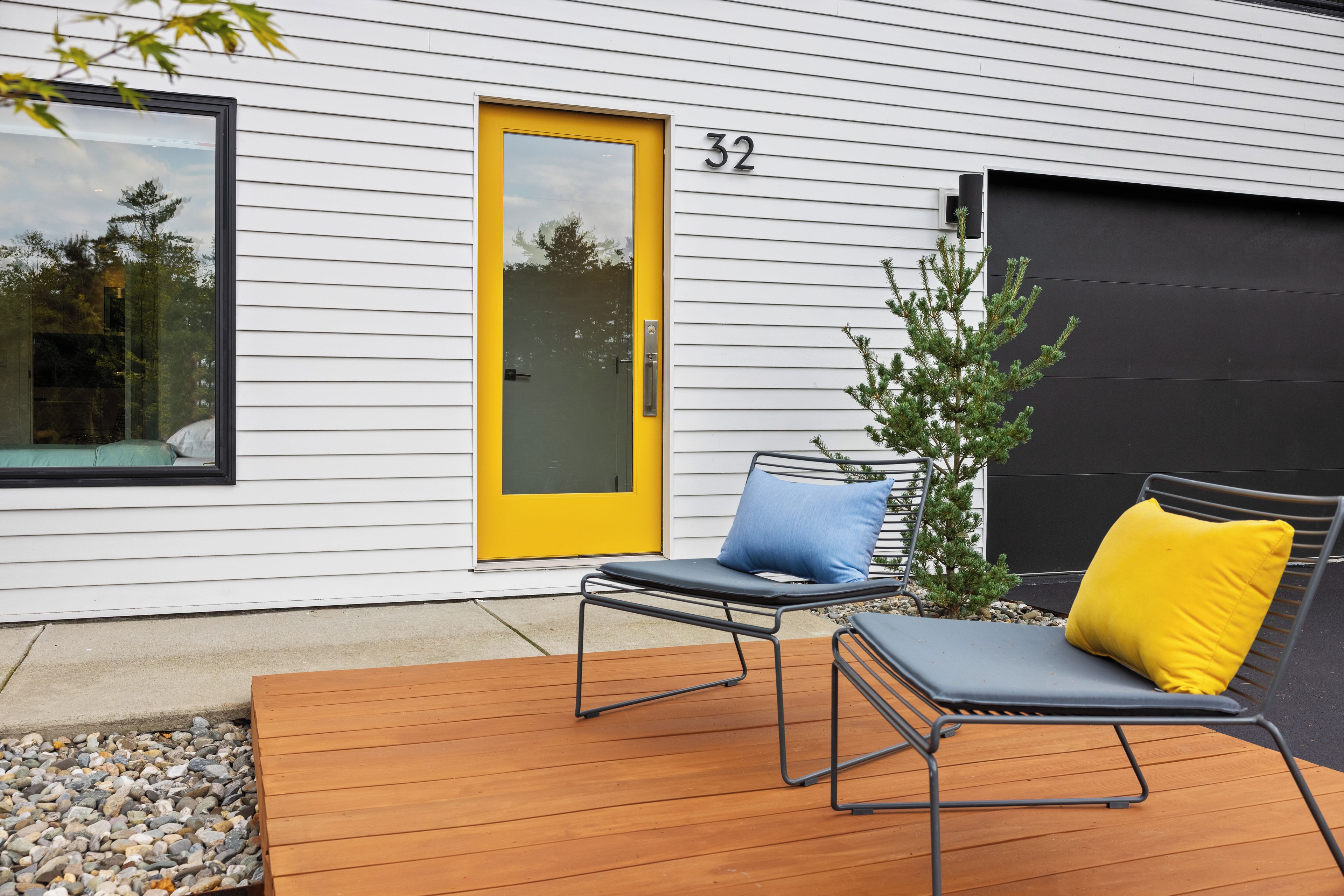Une maison moderne avec une porte jaune, le numéro 32, et une terrasse dotée de deux chaises longues et d’un petit arbre.