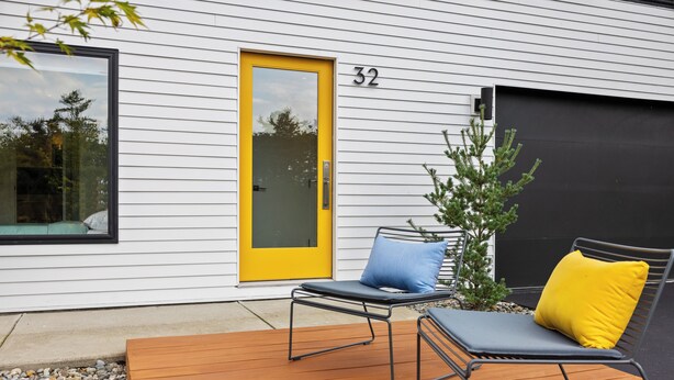Una casa moderna con una porta gialla, il numero 32, e una terrazza con due poltrone e un piccolo albero.