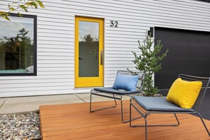 A modern house with a yellow door, number 32, and a deck with two lounge chairs and a small tree.