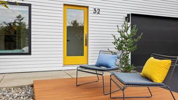 Une façade moderne avec une porte jaune, portant le numéro 32, et une terrasse en bois agrémentée de deux chaises longues et d’un petit arbre.