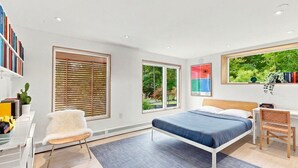 Una camera da letto moderna con un letto, una scrivania e una sedia. Grandi finestre offrono vista su un’area verde.