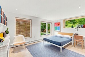 Quarto moderno com cama, escrivaninha e cadeira. Grandes janelas oferecem vista para o verde lá fora.