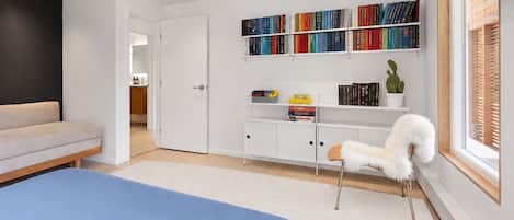 A bedroom with a bed, a white chair, a bookshelf, and a window.