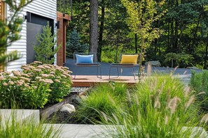 A patio area with outdoor seating, surrounded by lush greenery and a wooden deck. - Cozy 1BR Hanover Apartment | Walk to Dartmouth (Hanover)