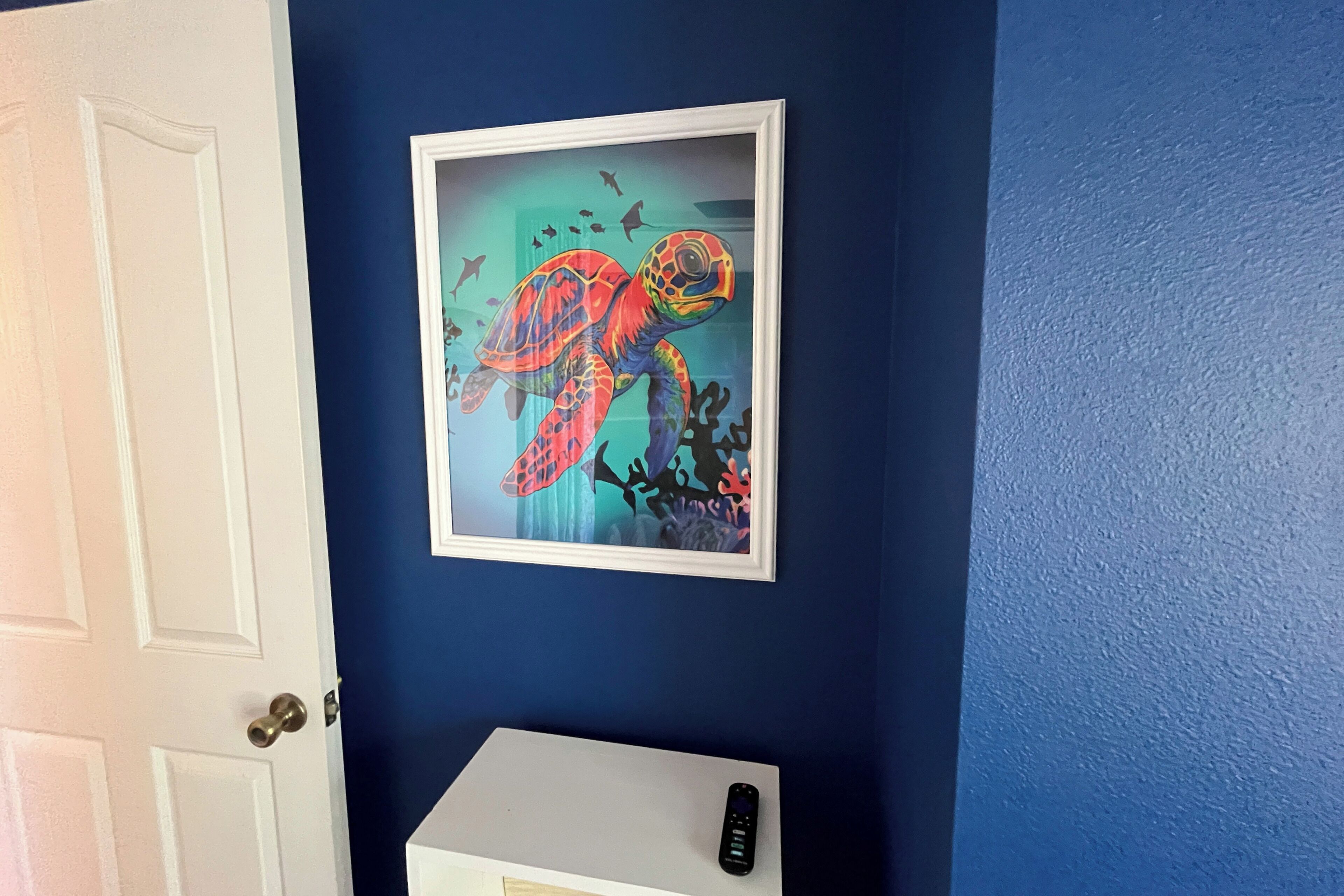 A framed artwork of a sea turtle on a blue wall, a white door, and a white cabinet with a remote control on it.