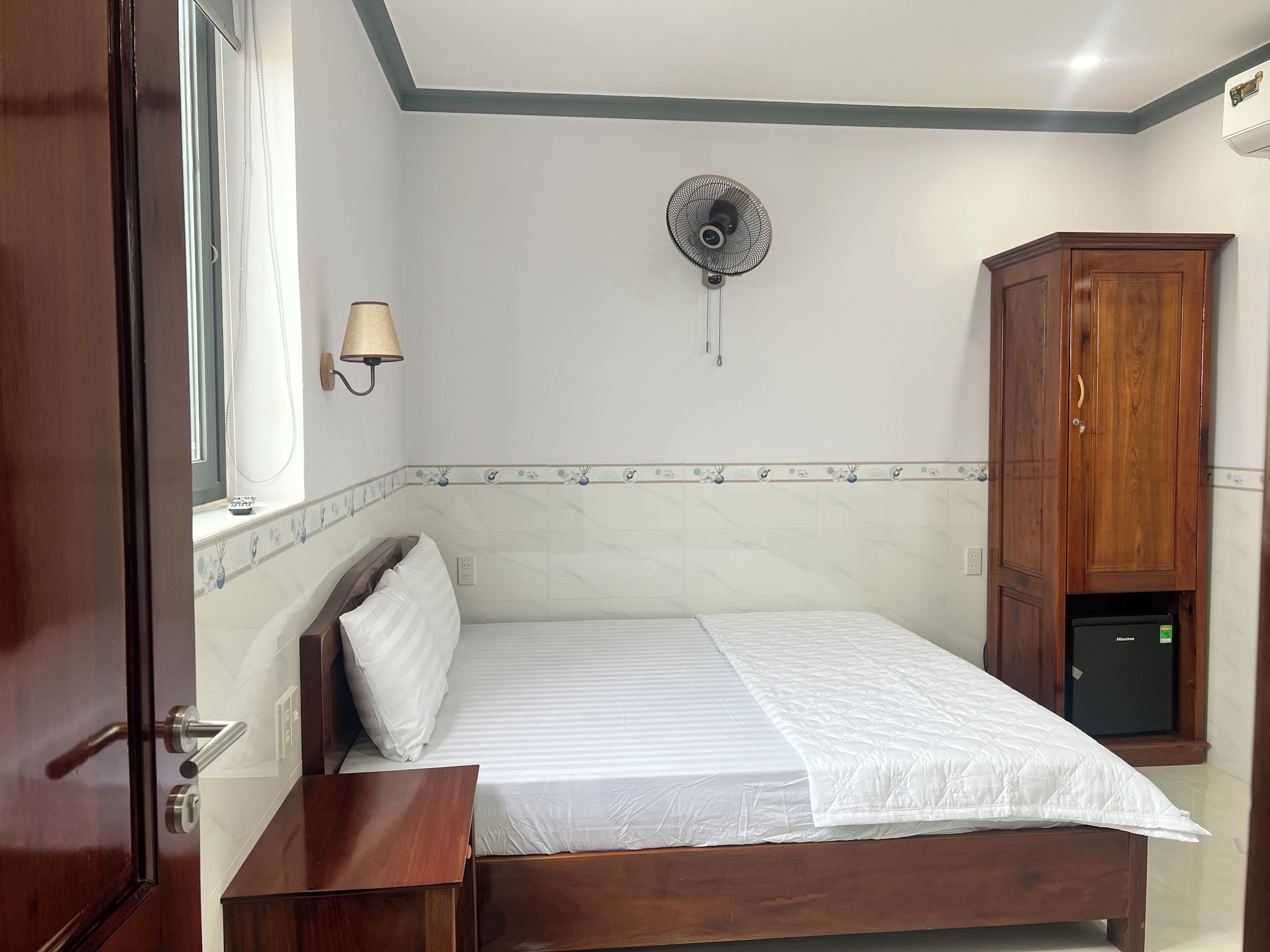 A bedroom with a bed, a bedside table, a wall-mounted fan, a cabinet, and a window with a curtain.