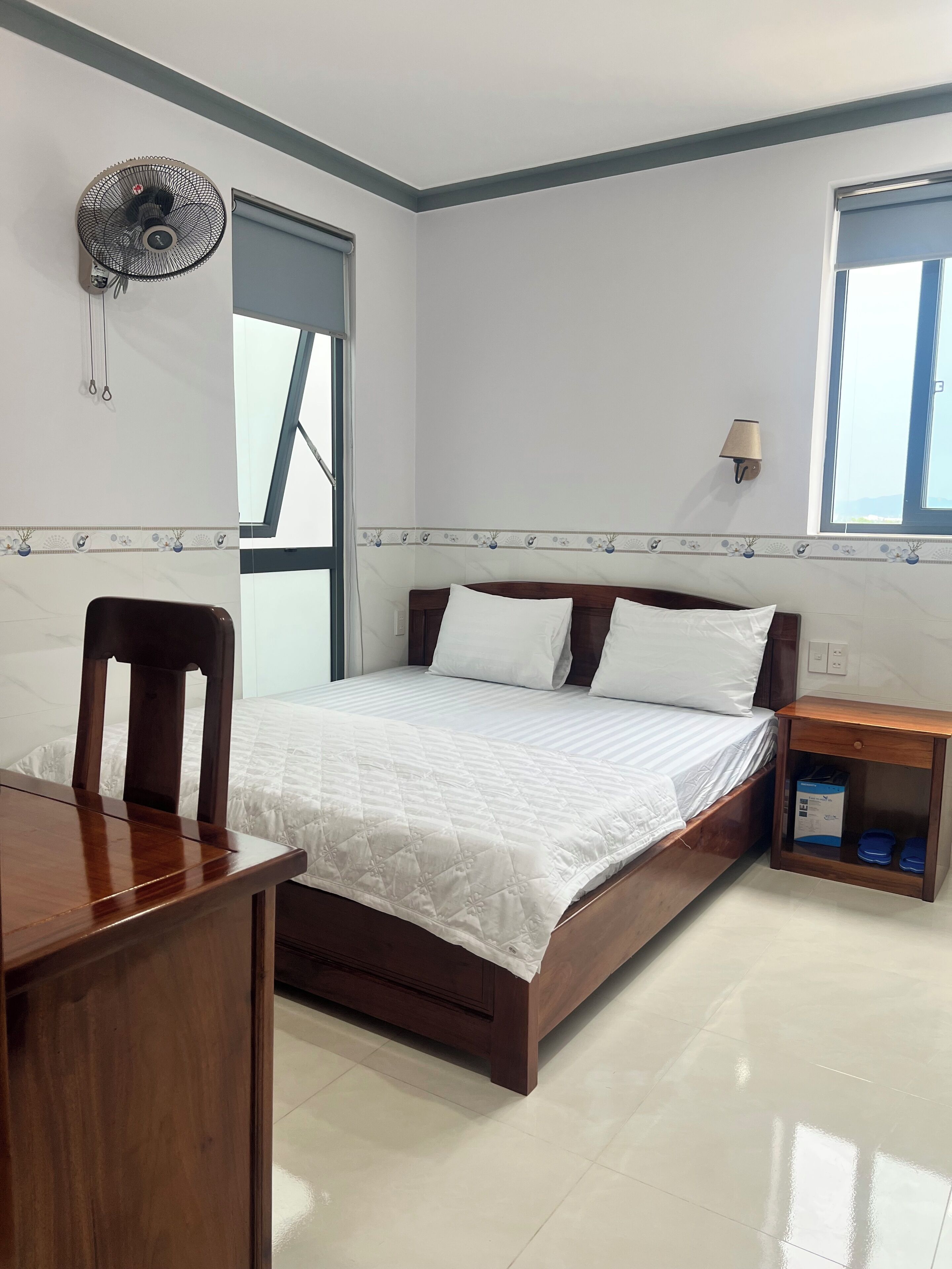 Une chambre à coucher comprenant un lit, une table de chevet en bois, un bureau, une chaise, un ventilateur fixé au mur et une fenêtre.