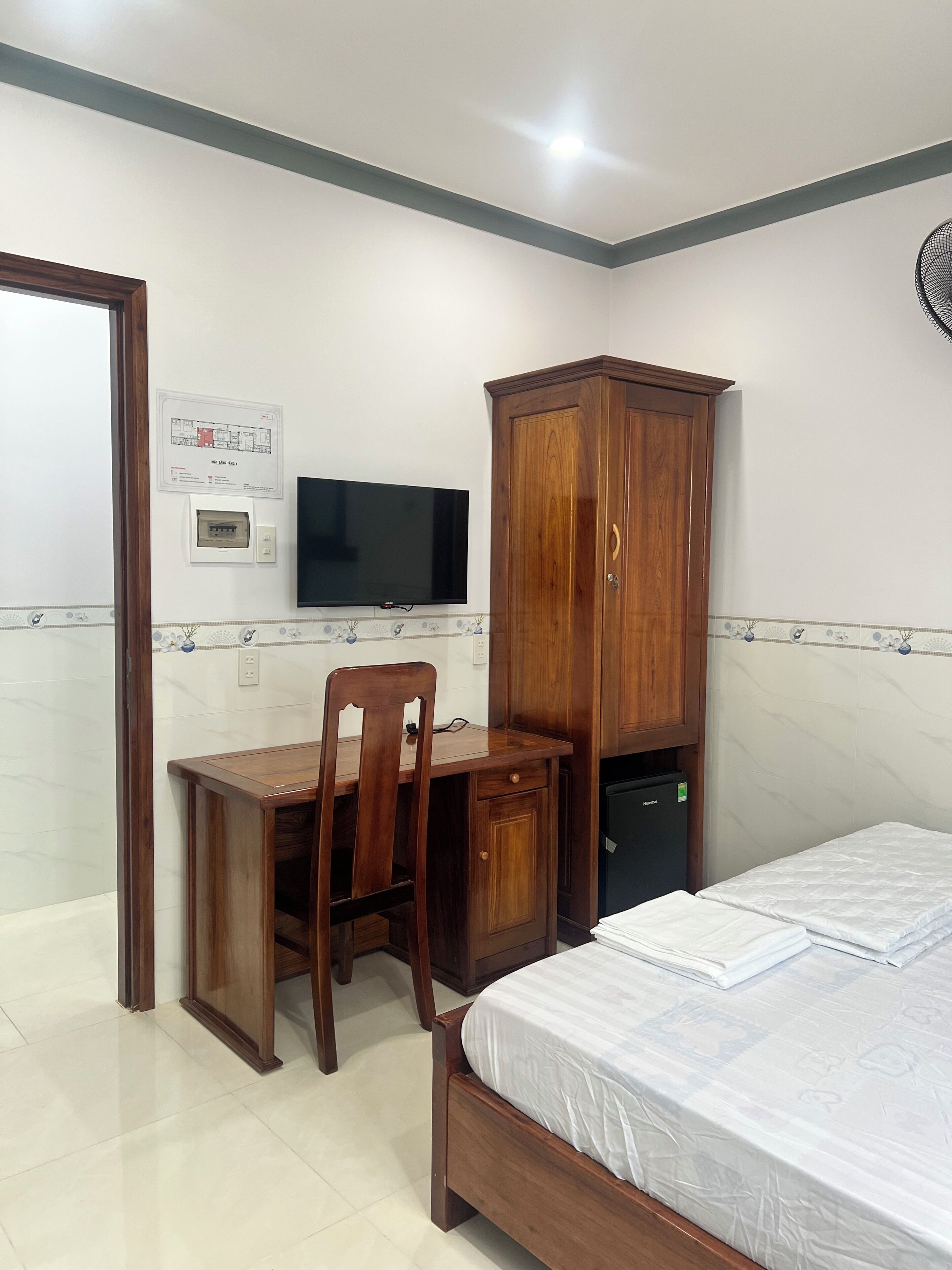 A bedroom with a bed, a desk with a chair, a wardrobe, a television, and a thermostat on the wall.
