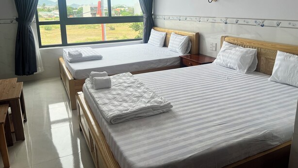 A hotel room with two beds, a wooden nightstand, a table, a window with curtains, a wall-mounted fan, and a lamp.