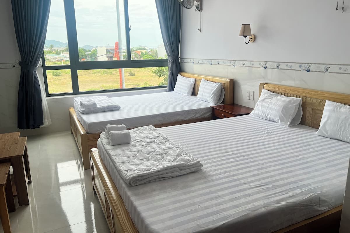 A hotel room with two beds, a wooden nightstand, a table, a window with curtains, a wall-mounted fan, and a lamp.