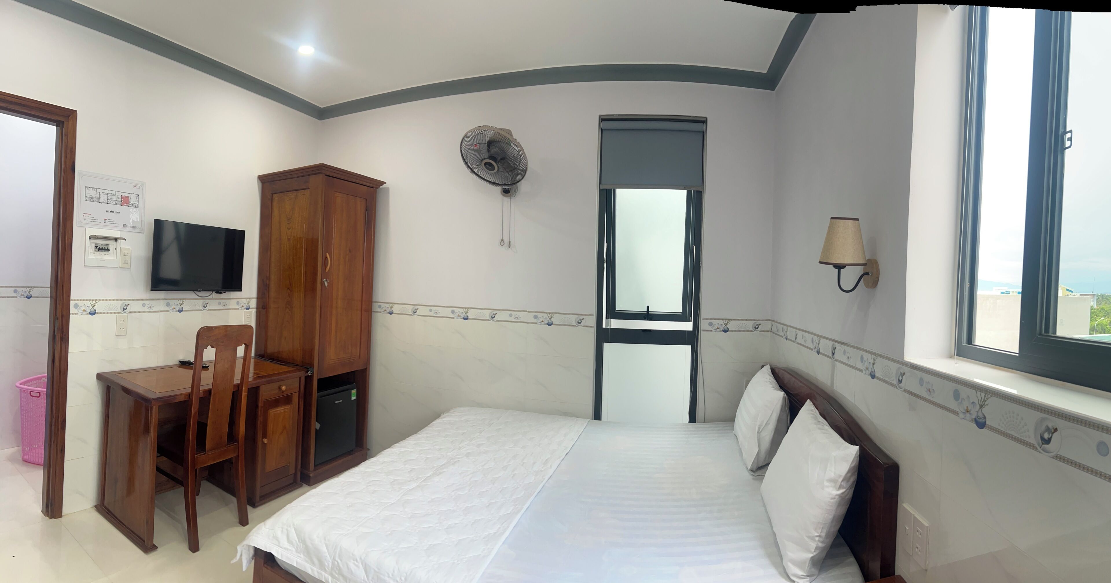 A bedroom with a bed, a desk with a chair, a wardrobe, a wall-mounted fan, a window with a view, and a wall-mounted lamp.