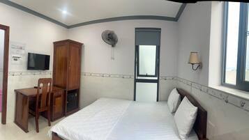 A bedroom with a bed, a desk with a chair, a wardrobe, a wall-mounted fan, a window with a view, and a wall-mounted lamp.