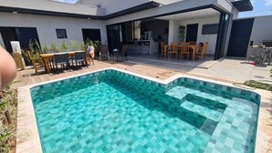 Una piscina moderna al aire libre con un diseño geométrico de azulejos, rodeada por una terraza con mobiliario exterior y una cocina visible al fondo.