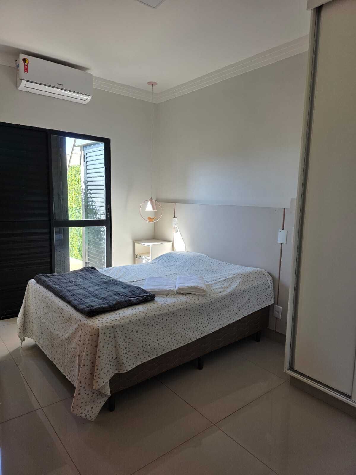 A bedroom with a bed, a bedside lamp, a window, and an air conditioning unit.