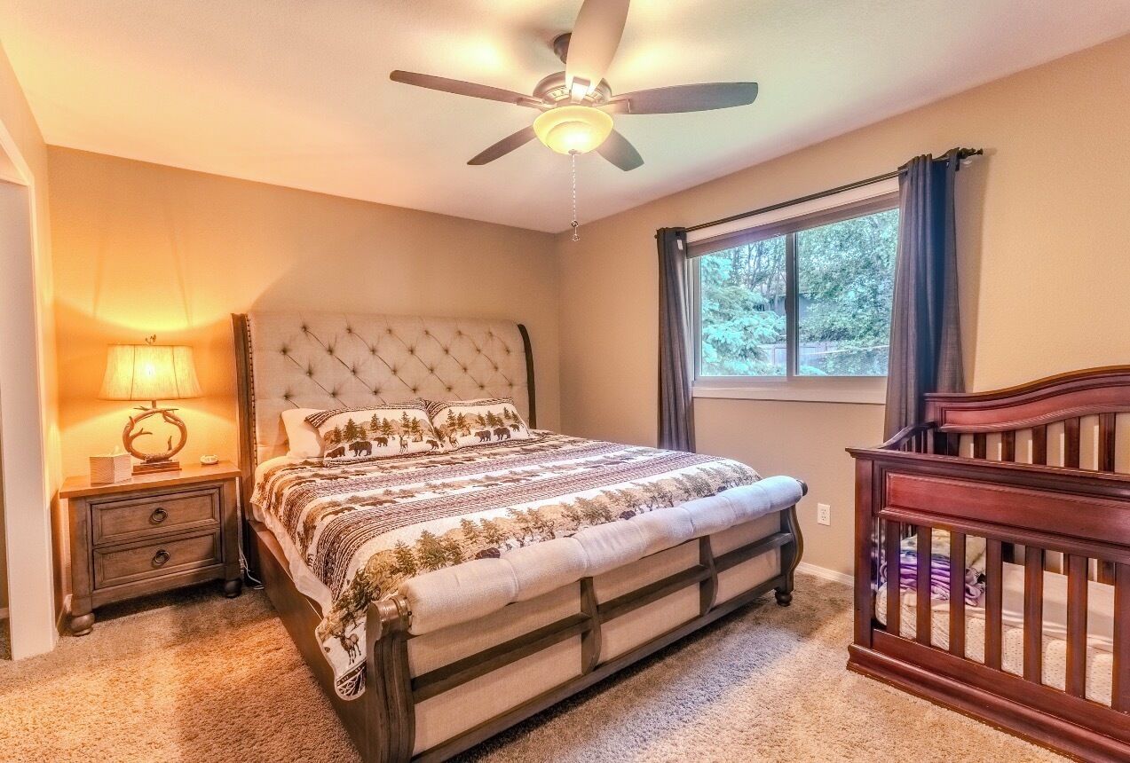 A bedroom with a bed, a nightstand with a lamp, a crib, a ceiling fan, and a window with curtains.