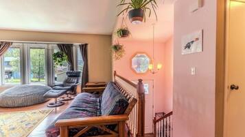 A cozy living room with a wooden sofa, a cushioned bench, and a chair. There are hanging plants and a hexagonal mirror on the wall.