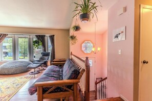 Una sala de estar acogedora con un sofá de madera, un banco acolchado y una silla. Hay plantas colgantes y un espejo hexagonal en la pared.