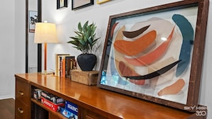 Una consola de madera con un cuadro abstracto enmarco, una planta en maceta, libros y juegos de mesa.