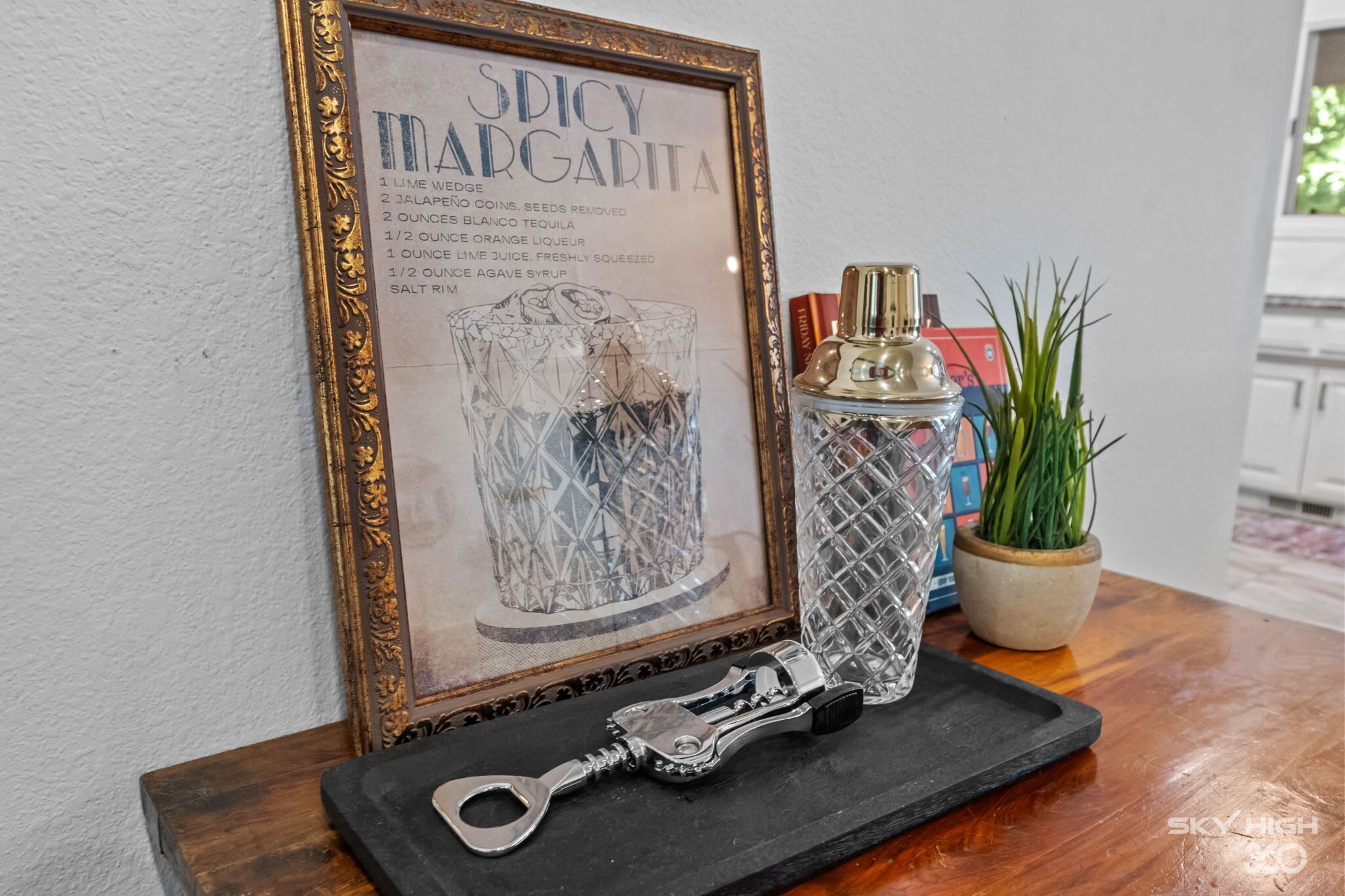 Tarjeta de receta enmarcada titulada 'Spicy Margarita' sobre una mesa de madera con un shaker de cócteles, una botella de licor y una planta en maceta.