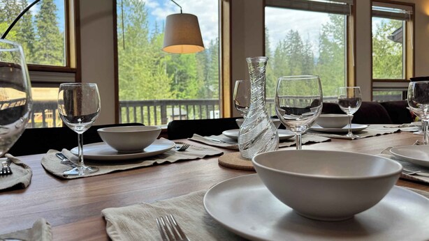 Une table à manger dressée avec de l’assiette blanche, des bols et des verres à vin, avec vue sur les arbres à travers de grandes fenêtres.