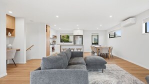 Ein modernes Wohnzimmer mit einem grauen Ecksofa, einem Essbereich mit Holztisch und Stühlen sowie einer Küche mit weißen Schränken und einem Fenster.