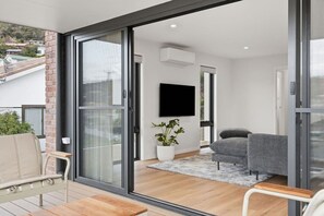 Ein modernes Wohnzimmer mit einer Schiebetür aus Glas, einem Flachbildfernseher, einem grauen Sofa, einem beige farbenen Sessel und einem Holztisch.