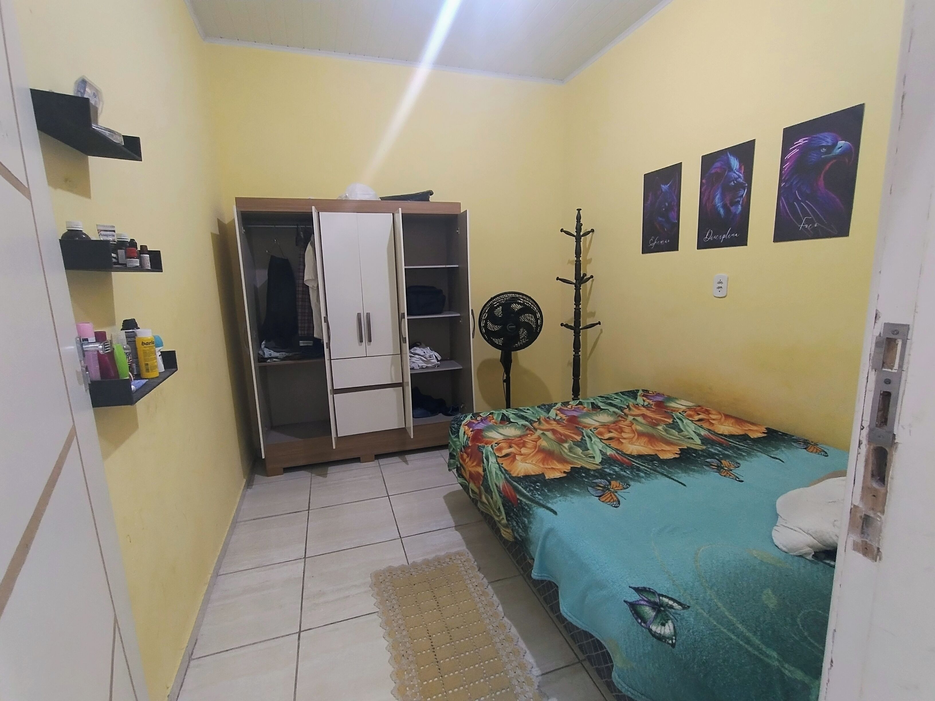 Een kleine slaapkamer met een bed, een kledingkast, een ventilator en een plank met toiletartikelen.