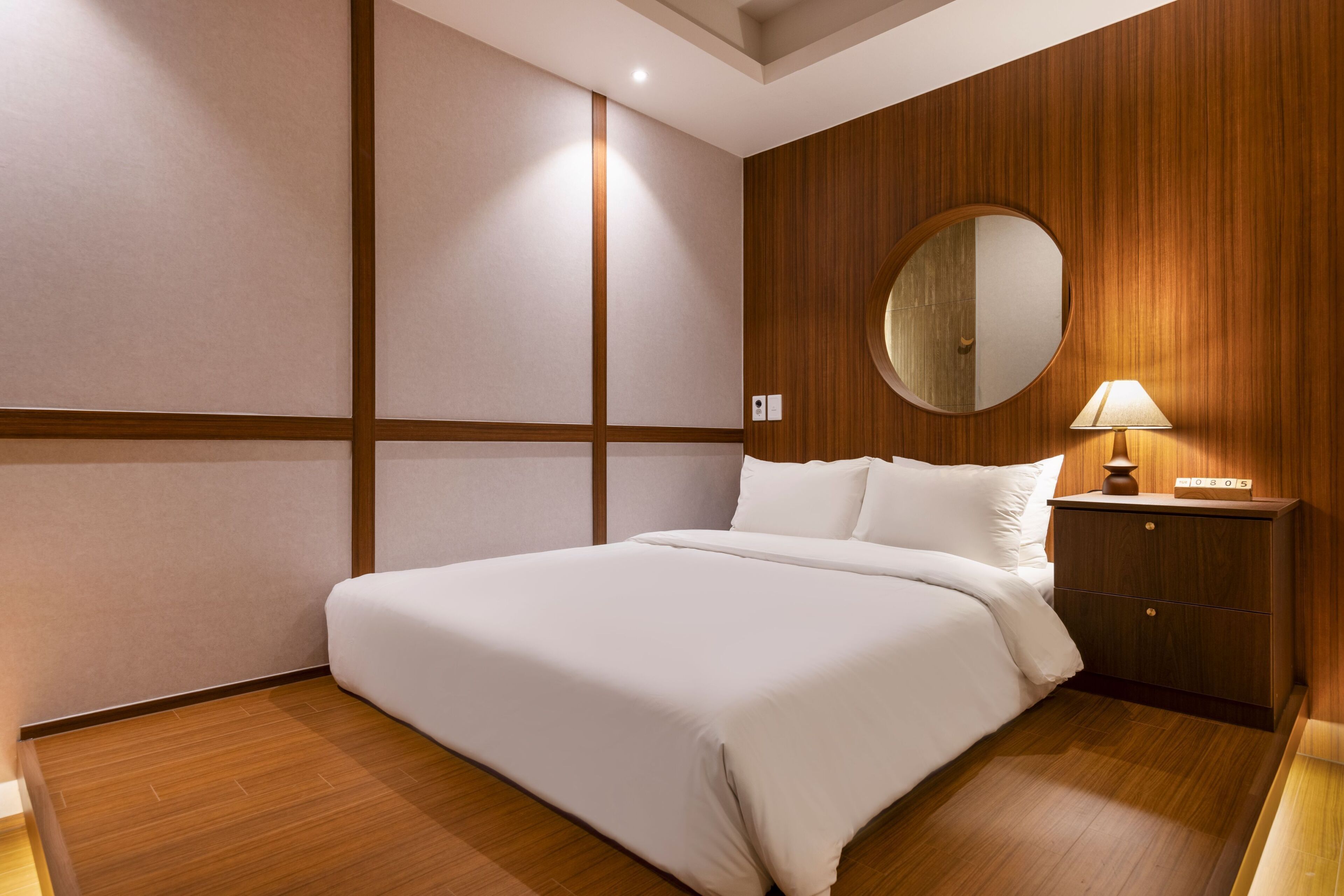 A hotel room with a large bed, a bedside table with a lamp, a wooden wall panel, and a round mirror.