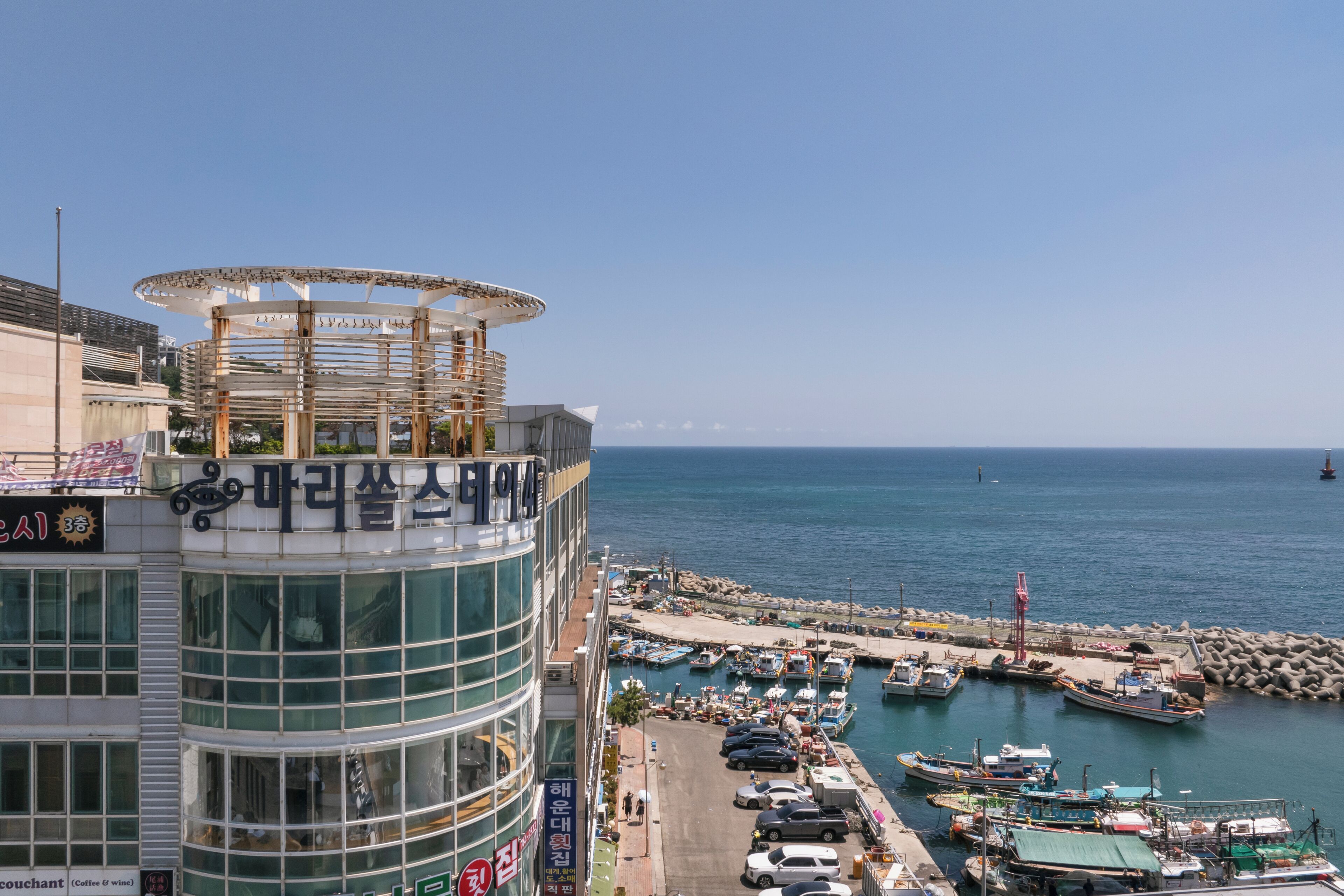 A coastal building with a curved glass facade, a rooftop terrace, and a view of a marina with boats.