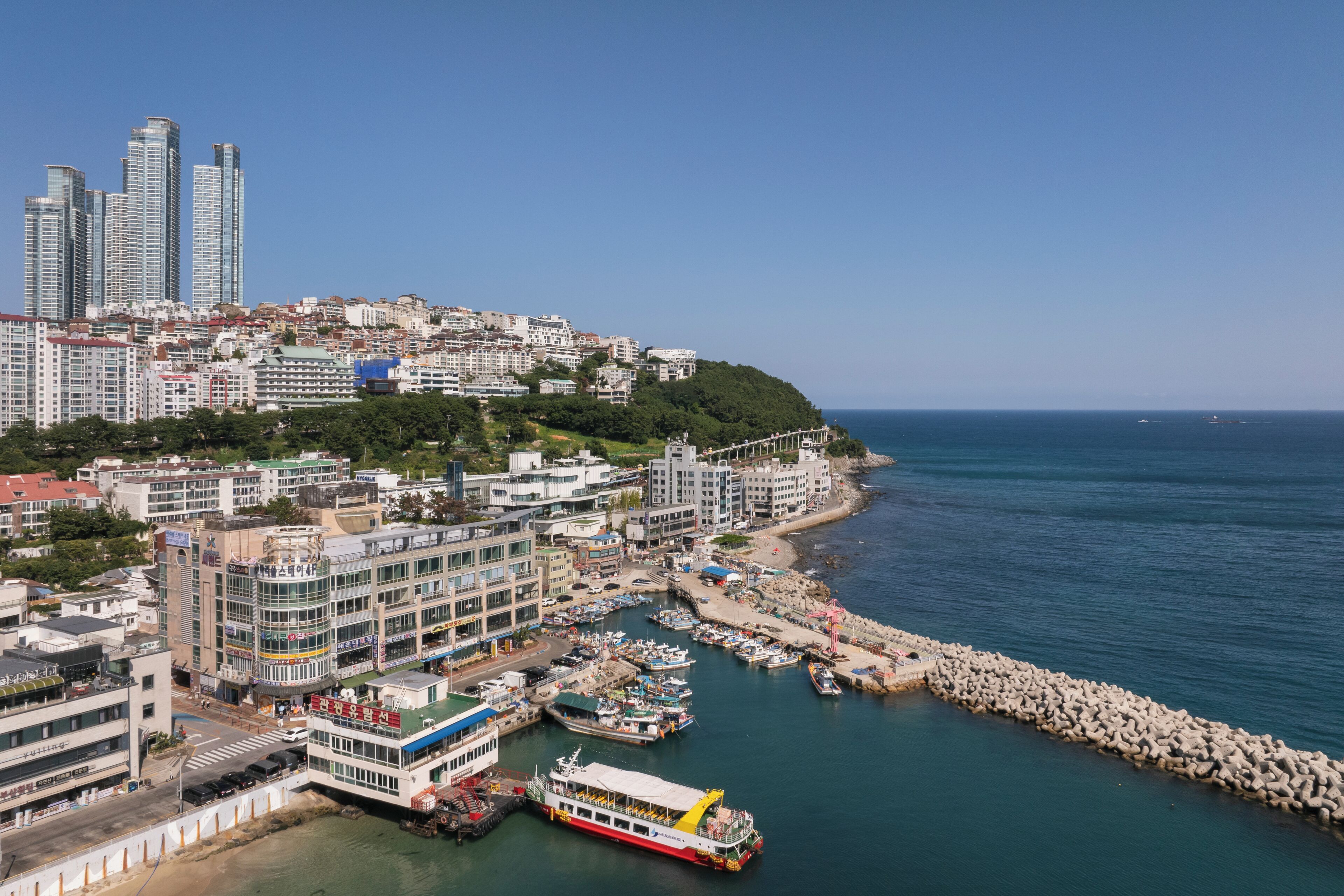 A coastal cityscape with a marina, high-rise buildings, and a clear view of the sea.
