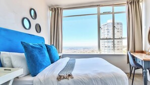 A hotel room with a large bed, a desk, and a view of a cityscape through a window. - Table Mountain Escape, Mountain and sea Views (Cape Town)