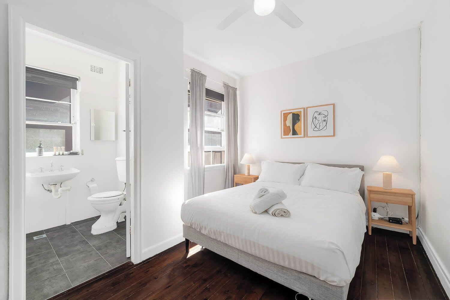 A bedroom with a bed, bedside lamps, a nightstand, a window with curtains, a bathroom with a toilet and sink, and a ceiling fan.