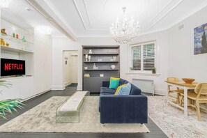 Ein Wohnzimmer mit einem dunkelblauen Sofa, einer Chandelüster, einem Bücherregal, einem Essbereich mit Tisch und Stühlen sowie einem Fernseher, der 'NETFLIX' anzeigt.