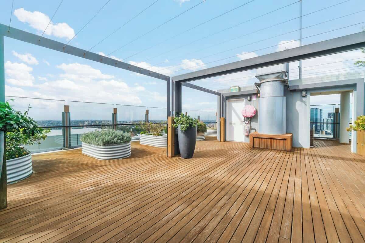 A rooftop terrace with wooden flooring, potted plants, a bench, and a view of the city skyline.