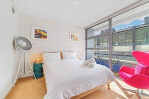 Ein modernes Hotelzimmer mit einem großen Bett, einer Stehlampe, einem hellen Fenster und einem rosa Sessel.