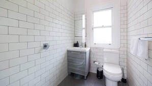 Ein Badezimmer mit weißen Fliesen, einer weißen Toilette, einem kleinen Waschbecken mit Spiegel und einem Fenster.