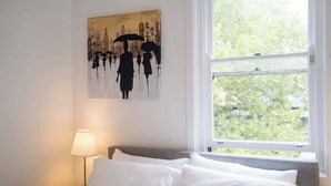 Ein Schlafzimmer mit einem Bett, weißen Kissen, einem Nachttisch mit Lampe und einem Bild von Personen mit Regenschirmen an der Wand.