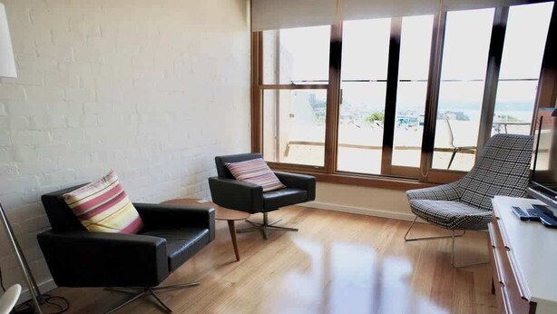 A modern living room with a black sofa, a patterned armchair, and a view of the city through large windows. - 1 bedroom apartment with amazing view (Paddington)