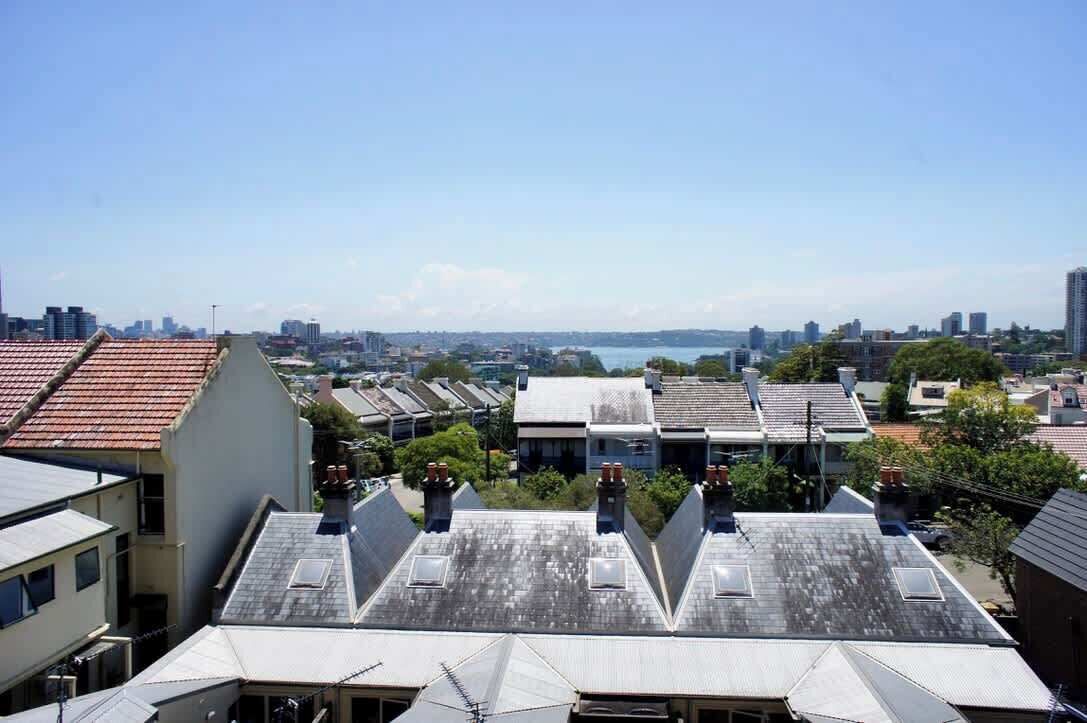 A residential area with multiple buildings, some with sloped roofs and chimneys, and a distant view of a city skyline and water body.