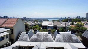A residential area with multiple buildings, some with sloped roofs and chimneys, and a distant view of a city skyline and water body. - 1 bedroom apartment with amazing view (Paddington)