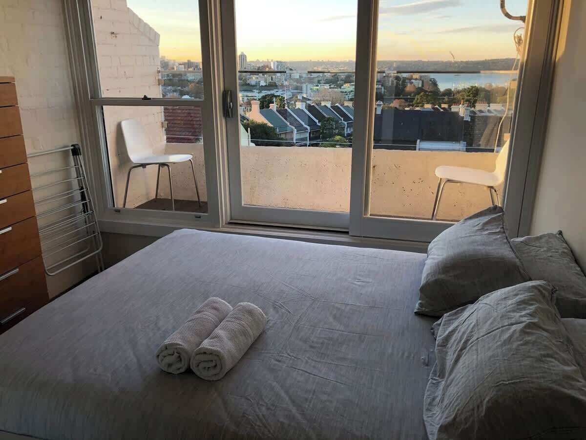 A bedroom with a bed, two pillows, a rolled towel, a chair, and a view of a cityscape through a large window.