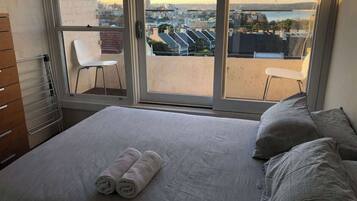 A bedroom with a bed, two pillows, a rolled towel, a chair, and a view of a cityscape through a large window.
