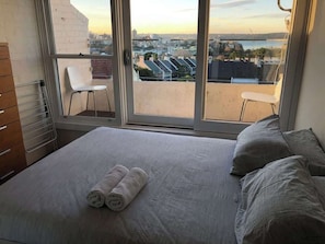 A bedroom with a bed, two pillows, a rolled towel, a chair, and a view of a cityscape through a large window. - 1 bedroom apartment with amazing view (Paddington)