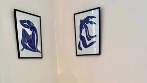 Huoneessa on vaaleanpunainen tuoli, puinen pöytä ja kaksi abstraktia taideteosta seinällä.