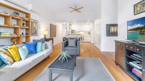 A modern living room with a sofa, coffee table, and bookshelf. There is a television on the right and a ceiling fan in the center. - Spacious 2 bedroom apt with parking (Surry Hills)