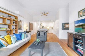 A modern living room with a sofa, coffee table, and bookshelf. There is a television on the right and a ceiling fan in the center. - Spacious 2 bedroom apt with parking (Surry Hills)