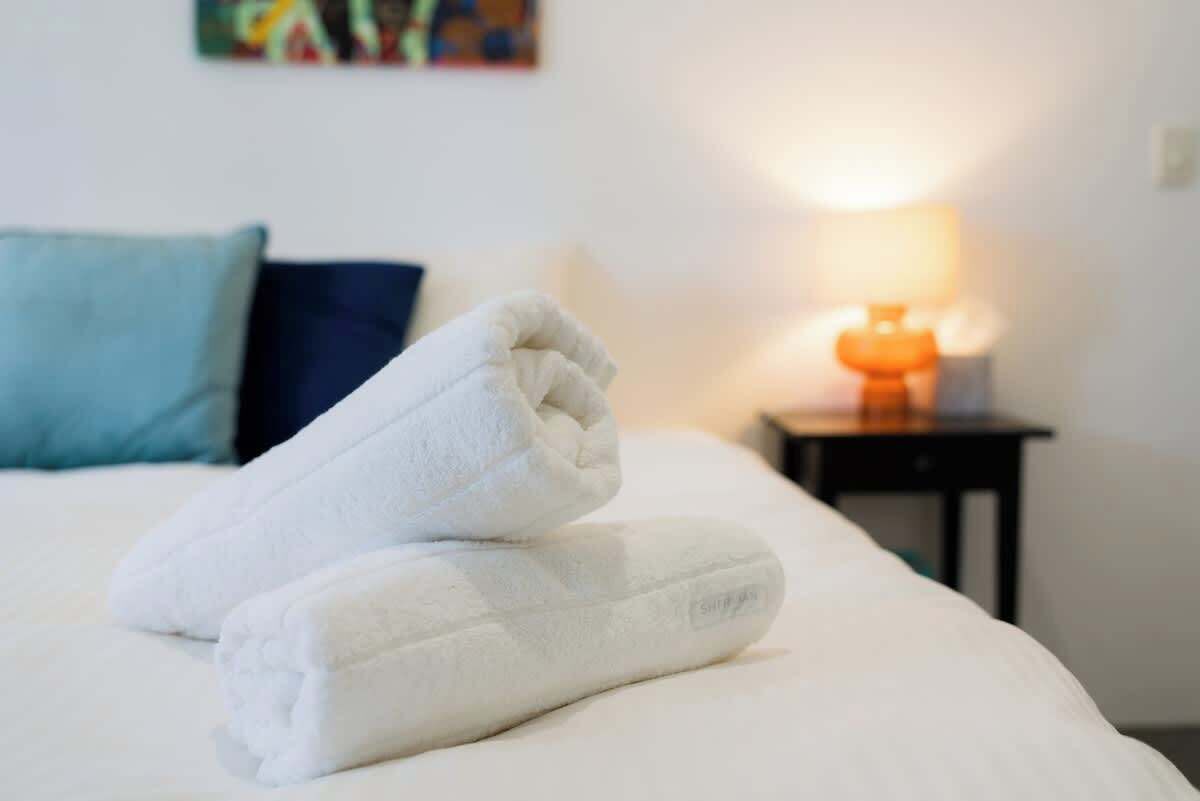 En pænt redt seng med hvide håndklæder, en blå pude og et lille bord med en lampe.